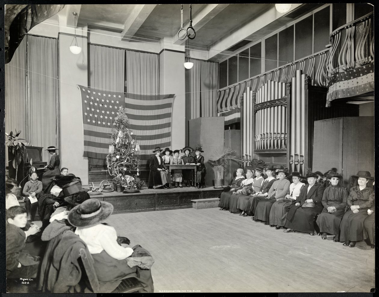 People in an Auditorium at the New York Association for the Blind with Christmas Tree and People Possibly Performing a Skit on Stage with Organ by Byron Company
