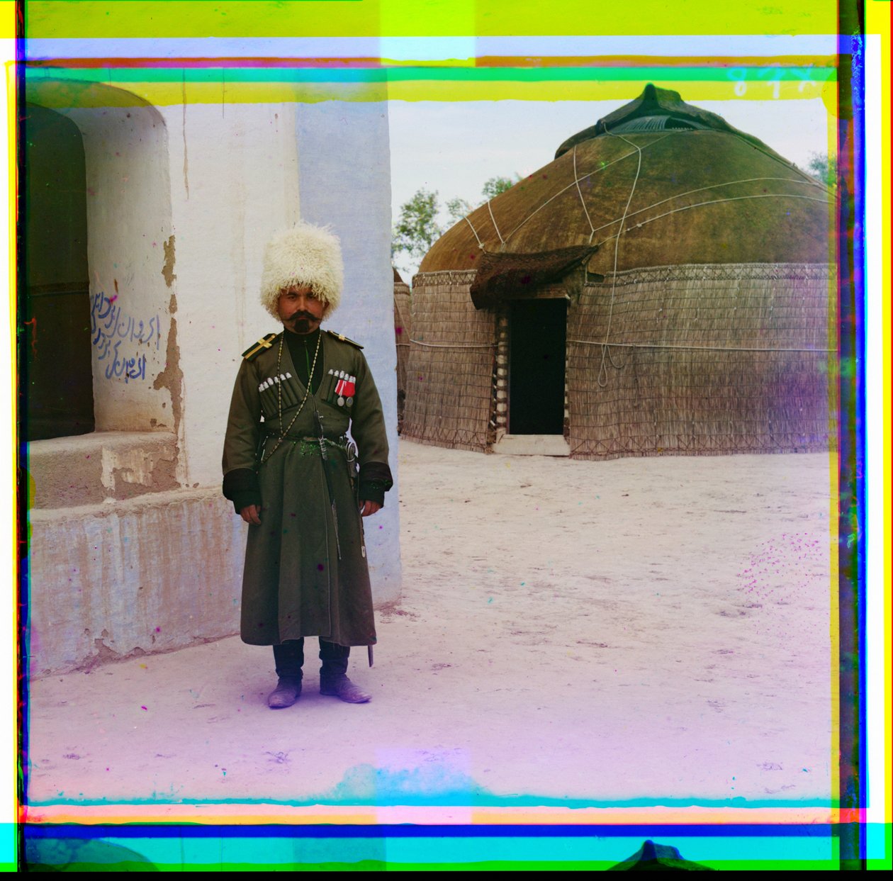 An Armed Man in Uniform Stands Next to Building with a Yurt in the Background, Russia, 1915 by Sergey Prokudin Gorsky